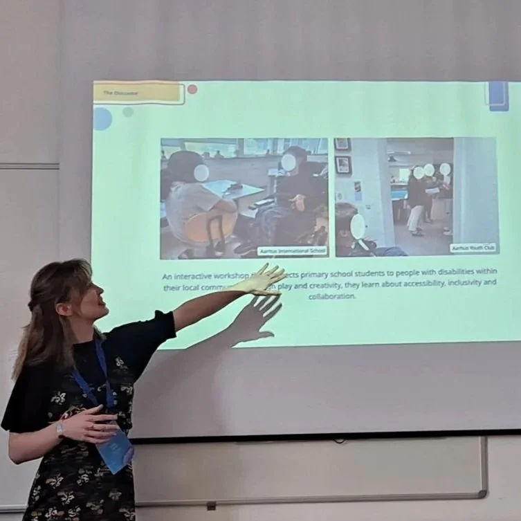 Sabhín is wearing a patterned jumpsuit and is gesturing in front of a projected slide. The slide shows two photos of site visits.