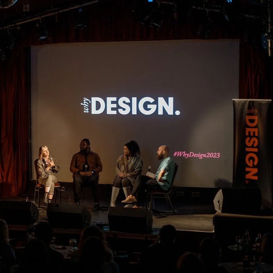 Four people are sitting on chairs on a stage. The spotlight is on a woman on the far left. A slide with the WhyDesign logo is on a black screen behind them. 