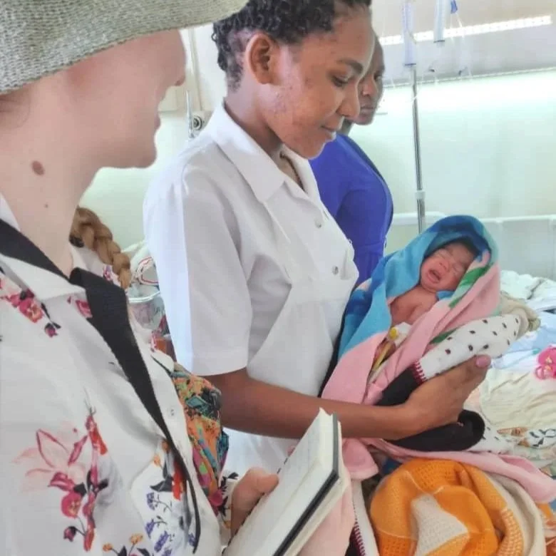 At the centre of the photo, a black woman in a nurse uniform is holding a premature baby swaddled in a pink blanket. Sabhín stands beside them, holding a notebook.