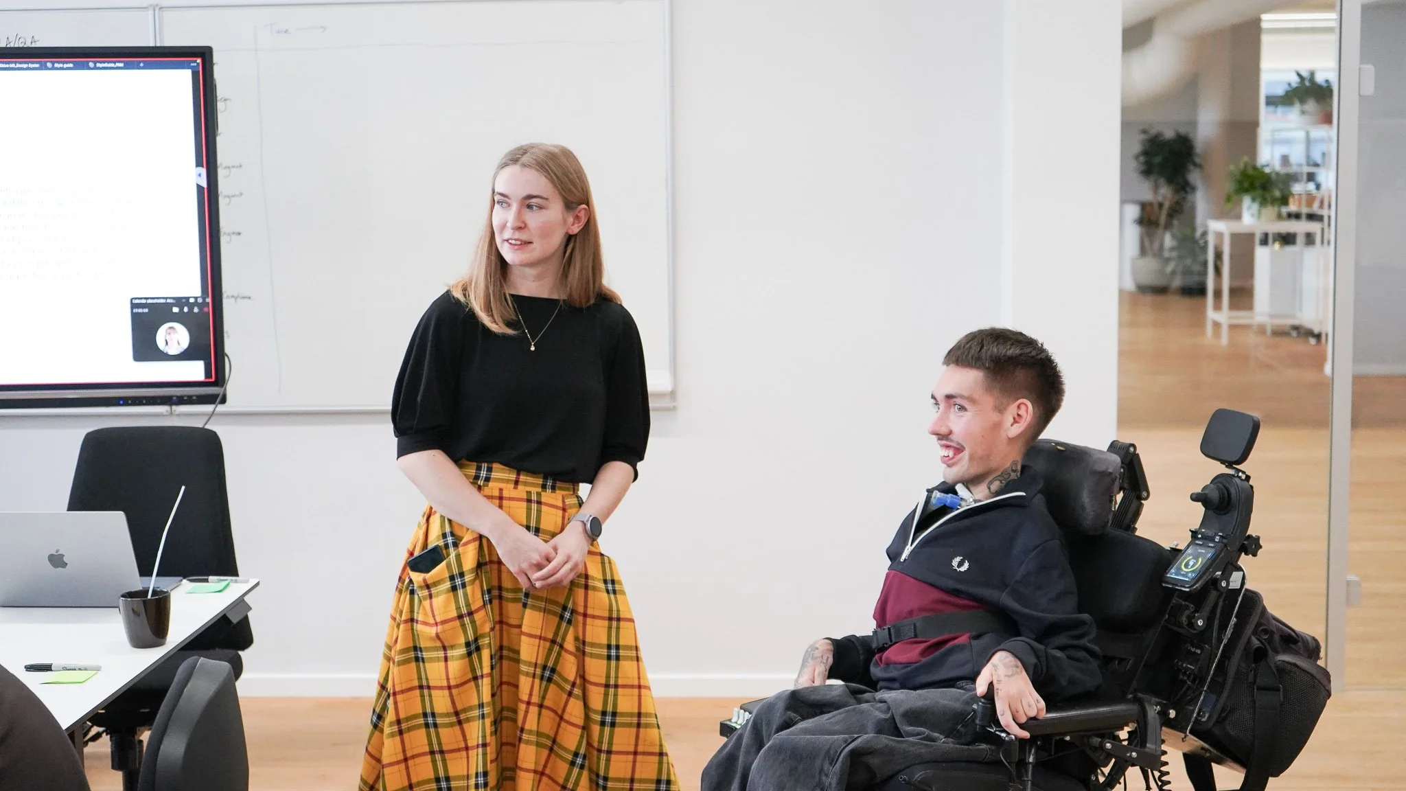 Sabhín stands to the left wearing a yellow skirt and a back top. William is on the right, smiling at the camera and wearing a navy and maroon t-shirt.