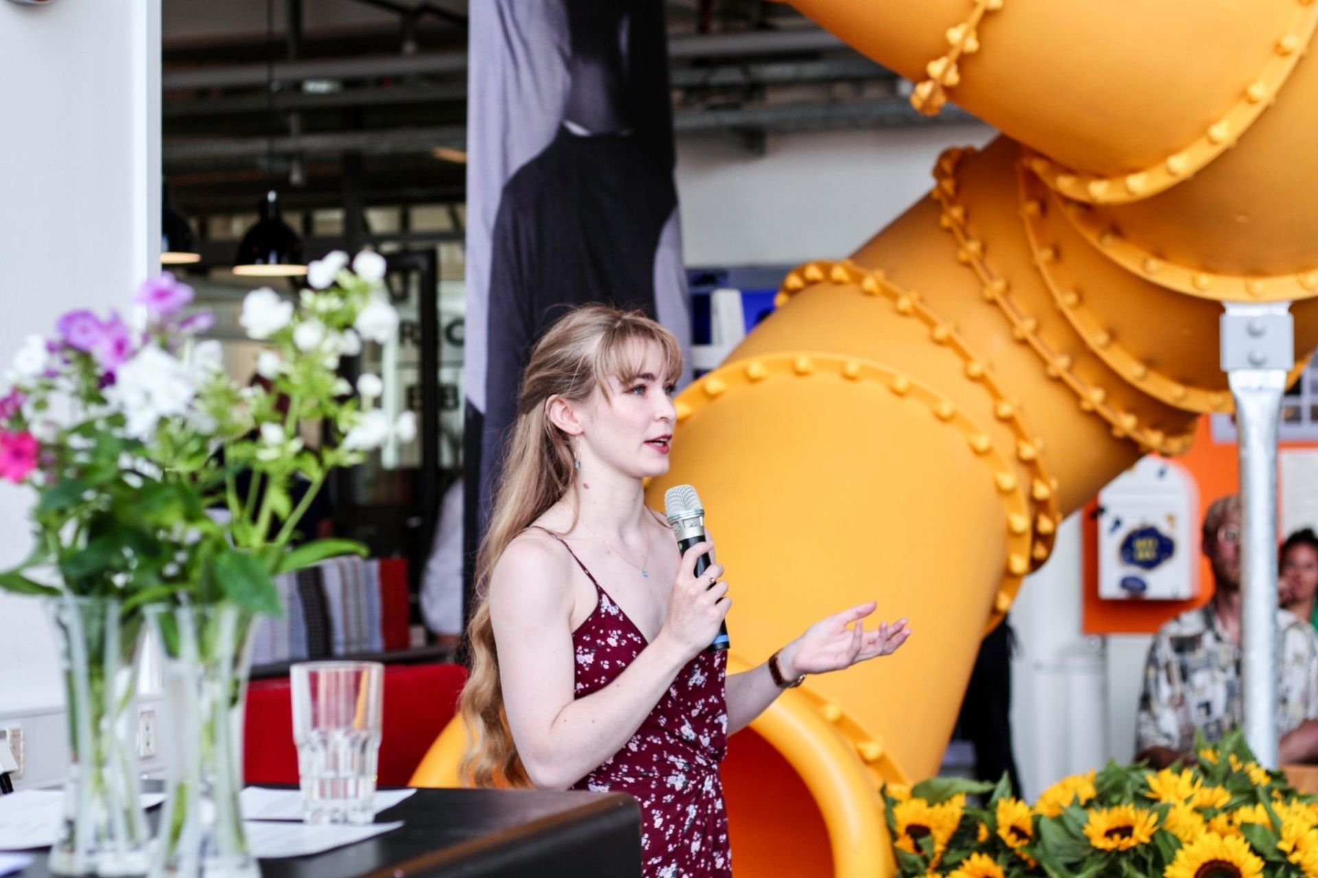 Sabhín is wearing a maroon dress, holding a microphone and gesturing as she talks.