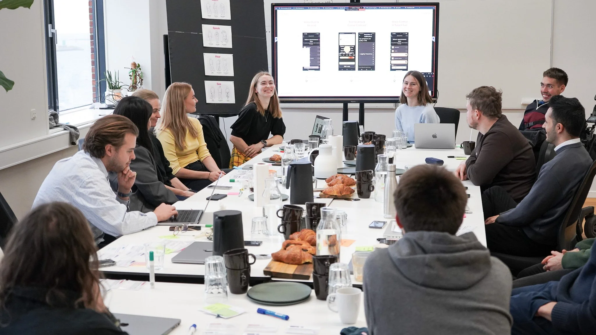 A group of employees sit around a white table and smiling. A large presentation screen showing some wireframes is positioned at the head of the table.