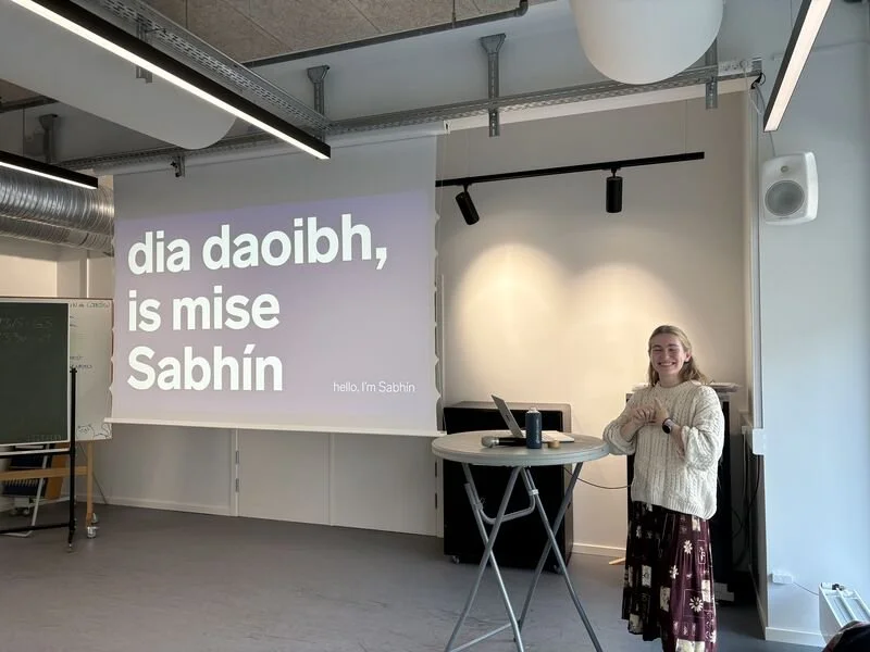 Sabhín is wearing a white jumper and maroon skirt. She is standing at a pedestal in front of a projection screen. On the screen are the words "dia daoibh, is mis Sabhín" (hello, I'm Sabhín).
