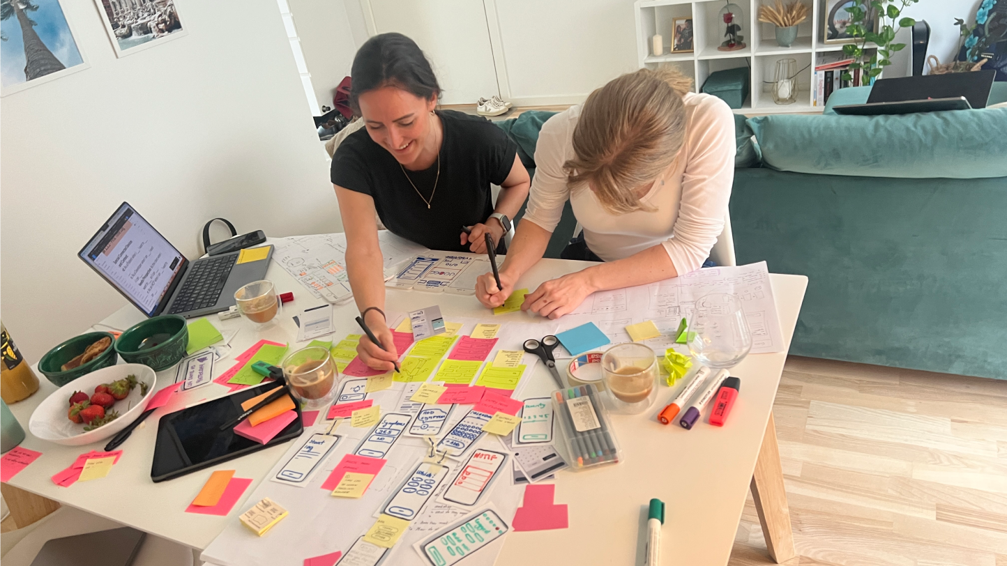 Two women are leaning over a table and drawing. The table is covered in post-it notes and wireframe sketches.