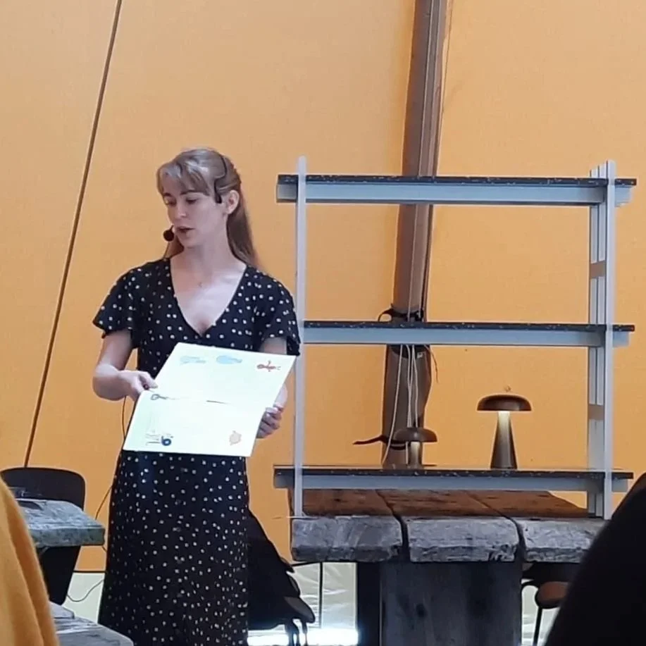 Sabhín is wearing a black dress and a handsfree microphone. She is holding a poster and speaking in front of an orange backdrop.