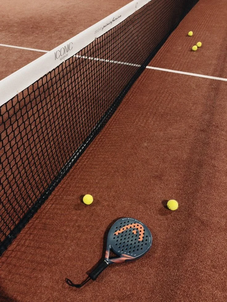 A tennis court with a black paddle and yellow tennis balls near the net.