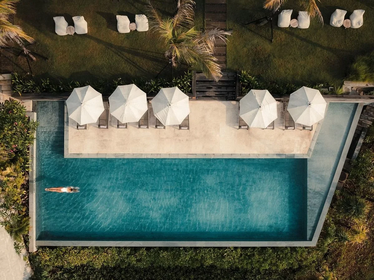 Aerial view of a swimming pool with a person relaxing on a float, surrounded by lounge chairs with umbrellas, palm trees, and greenery.