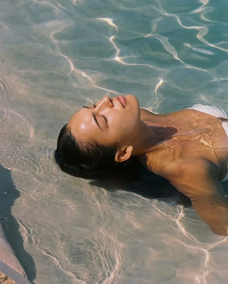 A woman with dark hair relaxing in shallow clear blue water with sunlight reflections.
