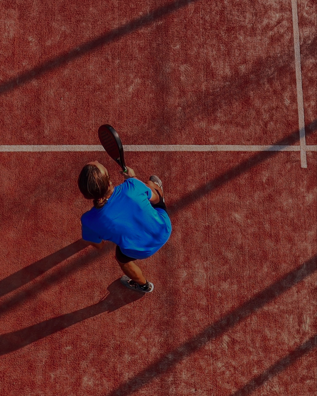 Top-down view of a person in a blue shirt holding a tennis racket on a red clay tennis court with long shadows.