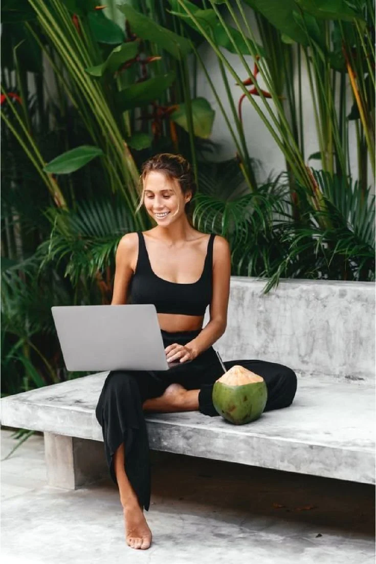 A young woman sitting on a concrete bench outdoors, working on a laptop, with a fresh coconut beside her, surrounded by lush green tropical plants.