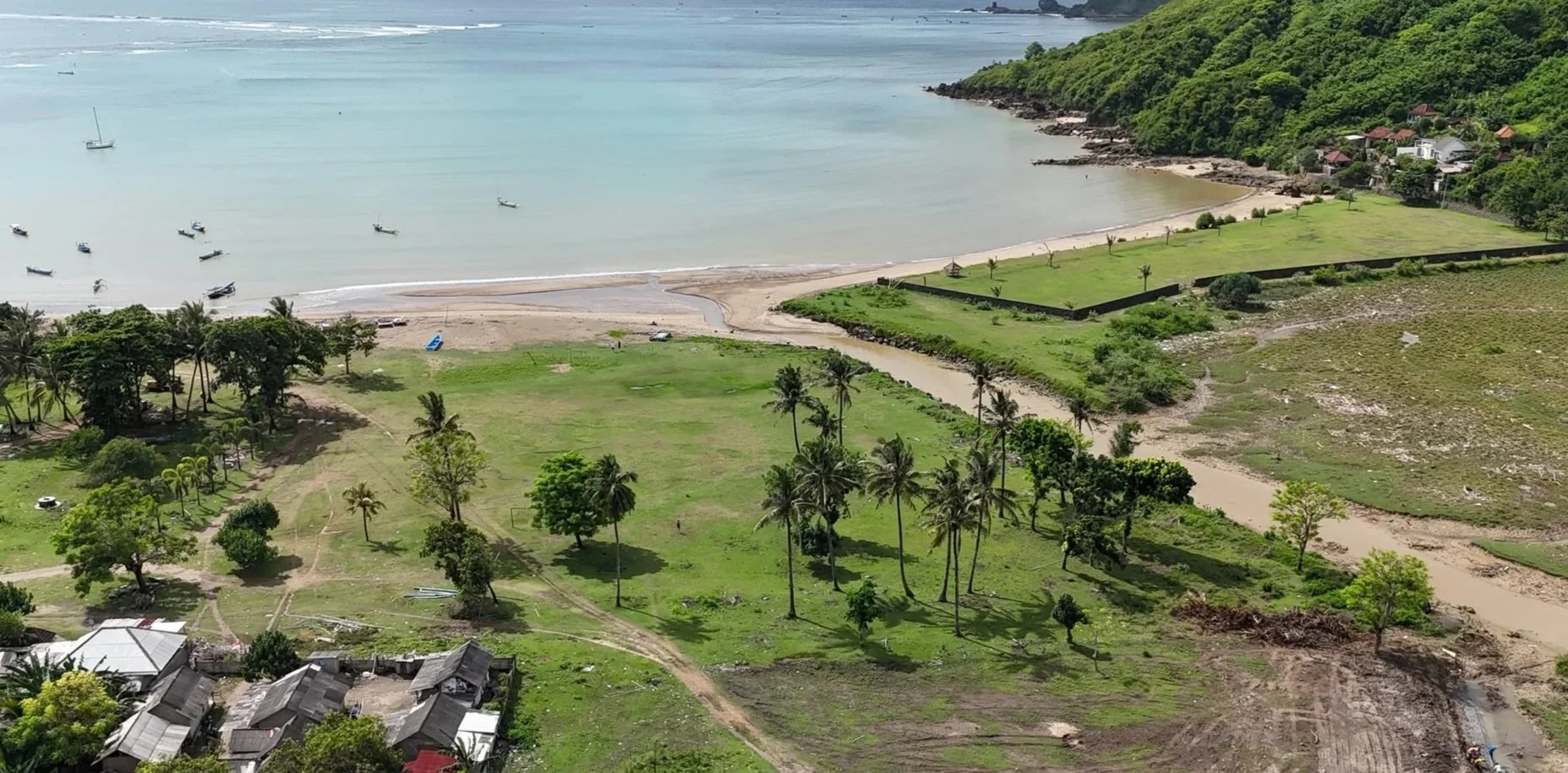 Aerial view of a coastal area with a beach, green grass, scattered palm trees, a river, and boats in the water.