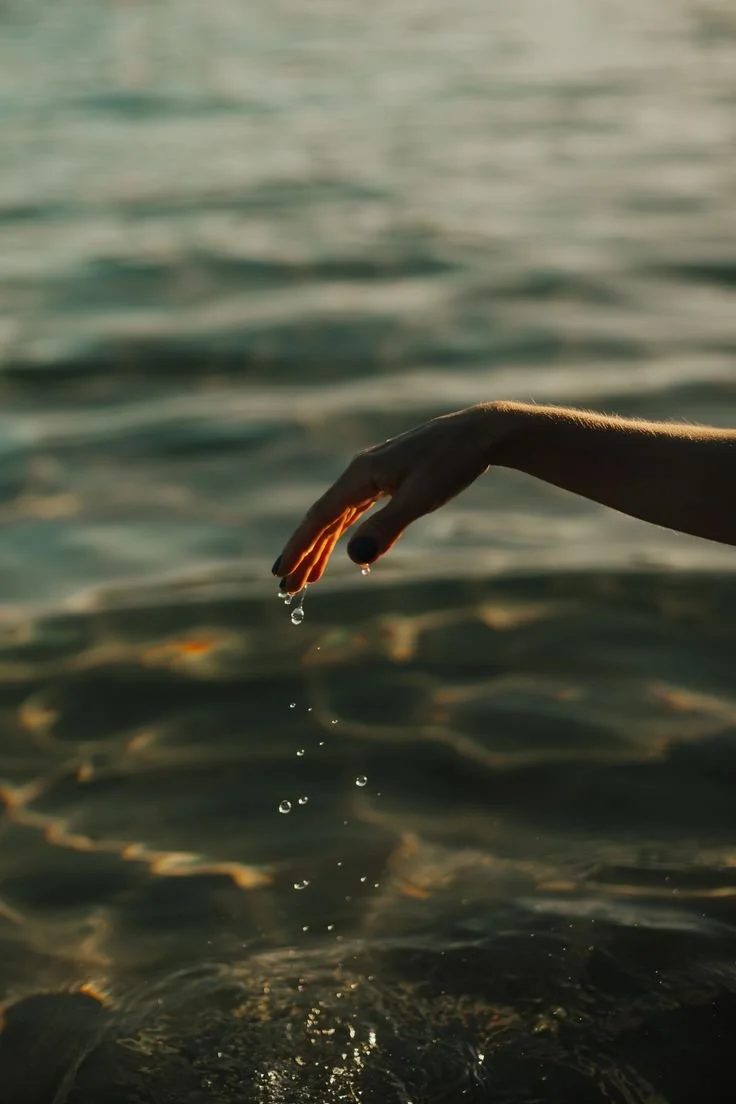 A hand dripping water over a body of water, with ripples and sunlight reflecting on the surface.