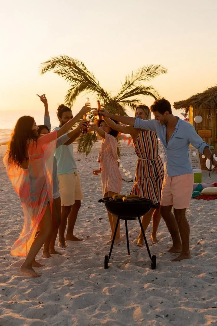 Group of friends having a toast with drinks on a beach at sunset, with palm trees and a thatched hut in the background.