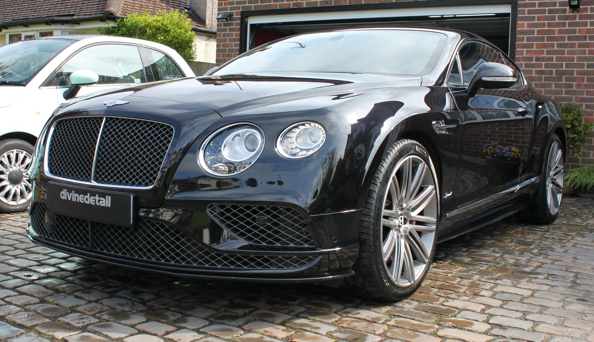 Black Bentley Continental GT parked on cobblestone driveway with white car and brick house in background.