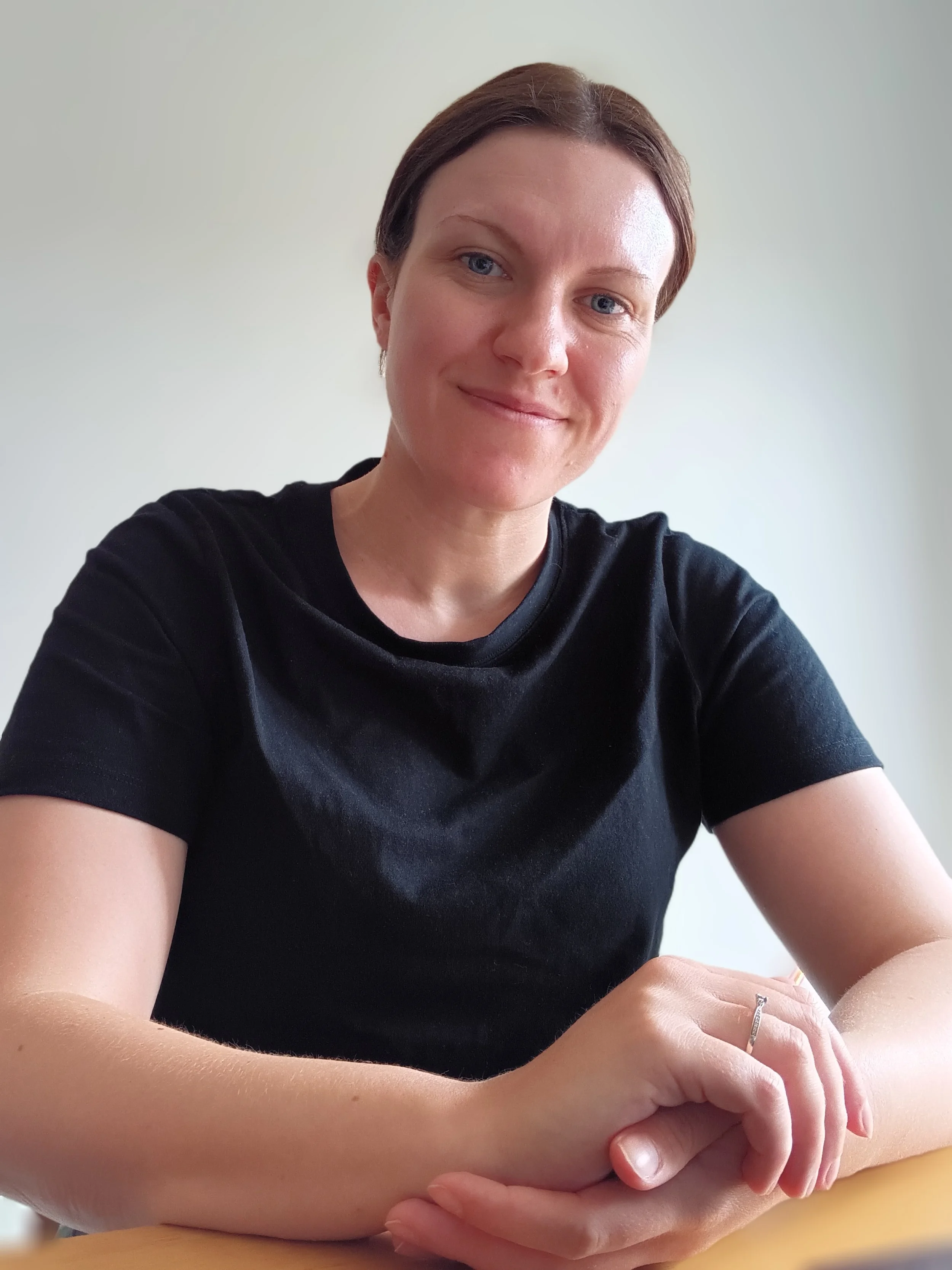 A woman with short brown hair wearing a black shirt, sitting at a table with her hands clasped, smiling softly, against a plain off-white background.