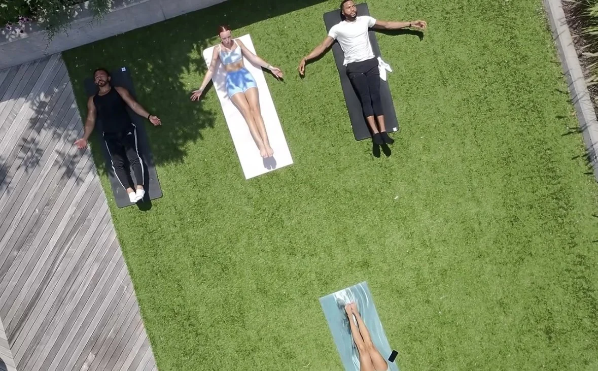Three people relaxing on yoga mats outdoors on a grassy area, with a fourth person lounging on a towel.