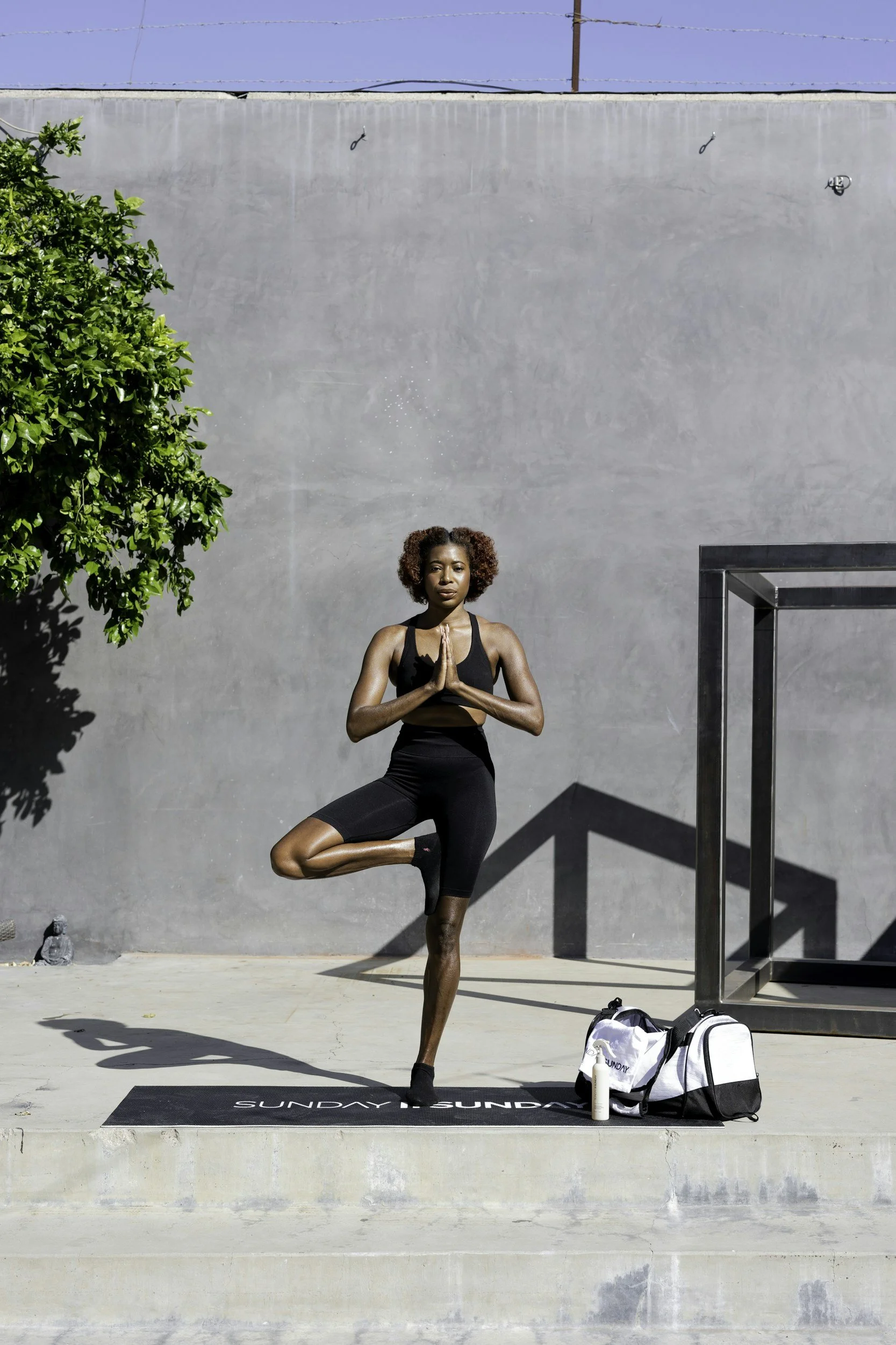 Woman practicing yoga in outdoor gym area balanced on one foot with palms pressed together, wearing black activewear, with yoga mat, water bottle, and gym bag nearby, sunny day with clear sky.