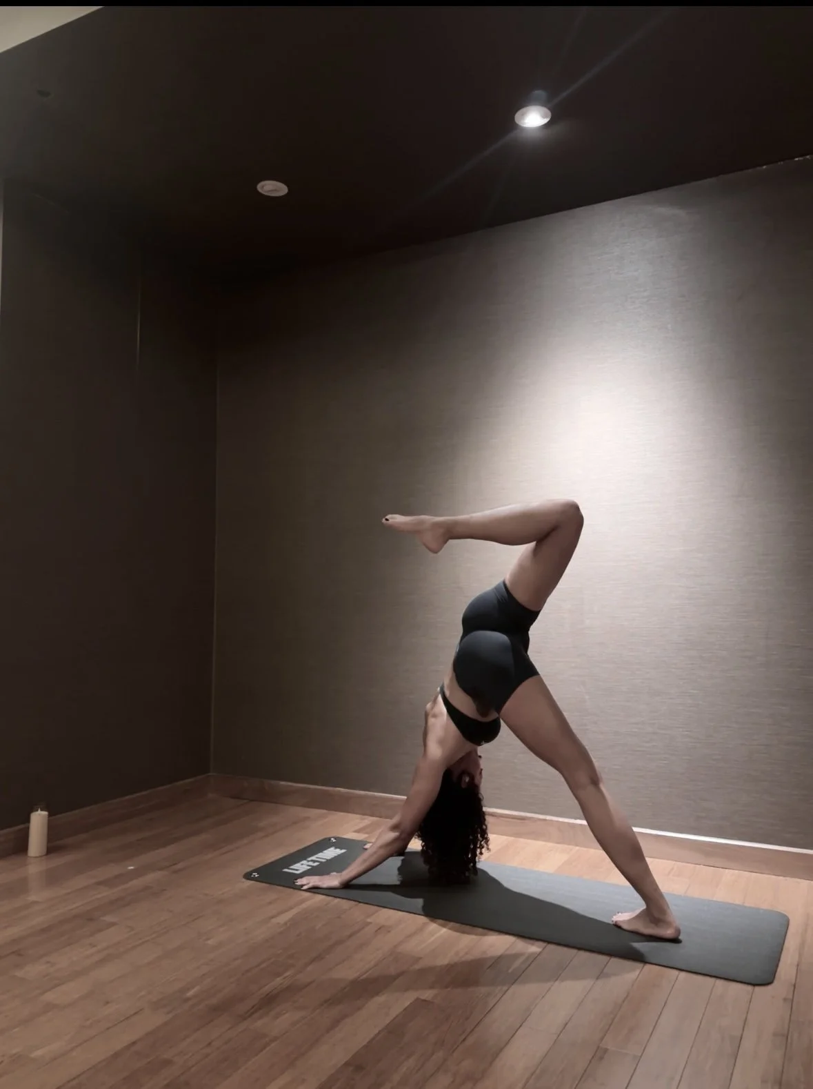 A woman practicing yoga in a dimly lit room, performing a downward dog pose with one leg lifted.