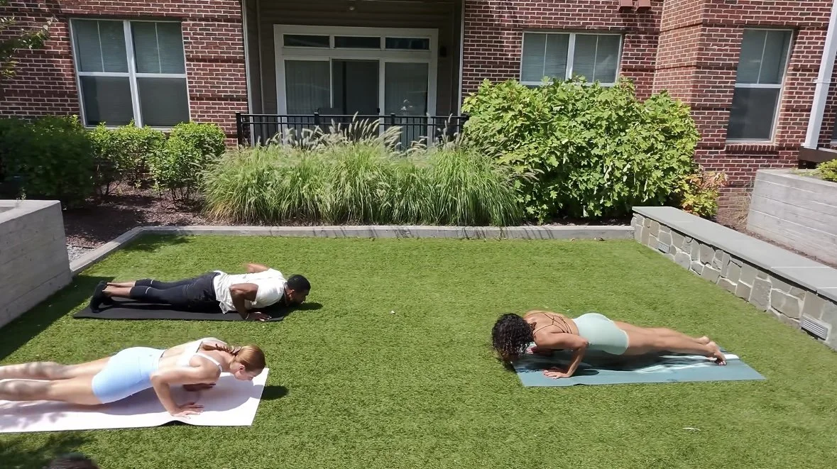Three people practicing yoga outdoors on a grassy area with a brick building and green bushes in the background. They are in different yoga poses on mats.