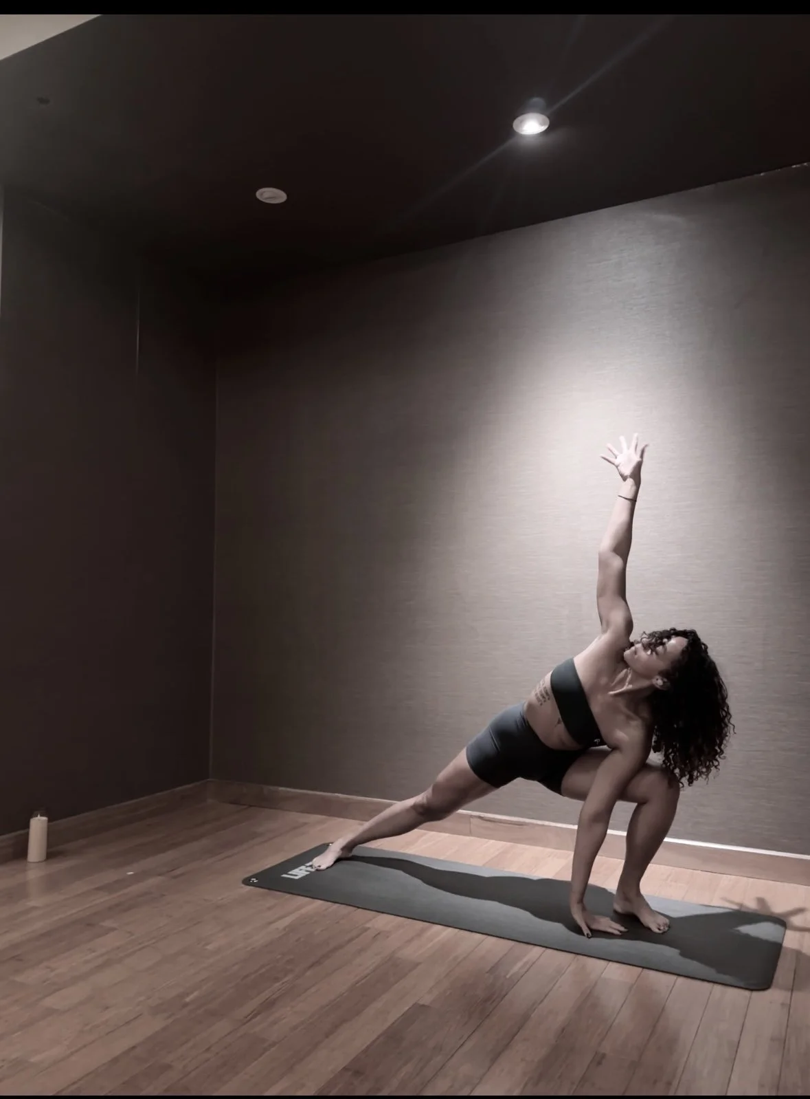 Woman practicing yoga in a dimly lit room, wearing workout clothes, on a yoga mat, with a candle in the corner.