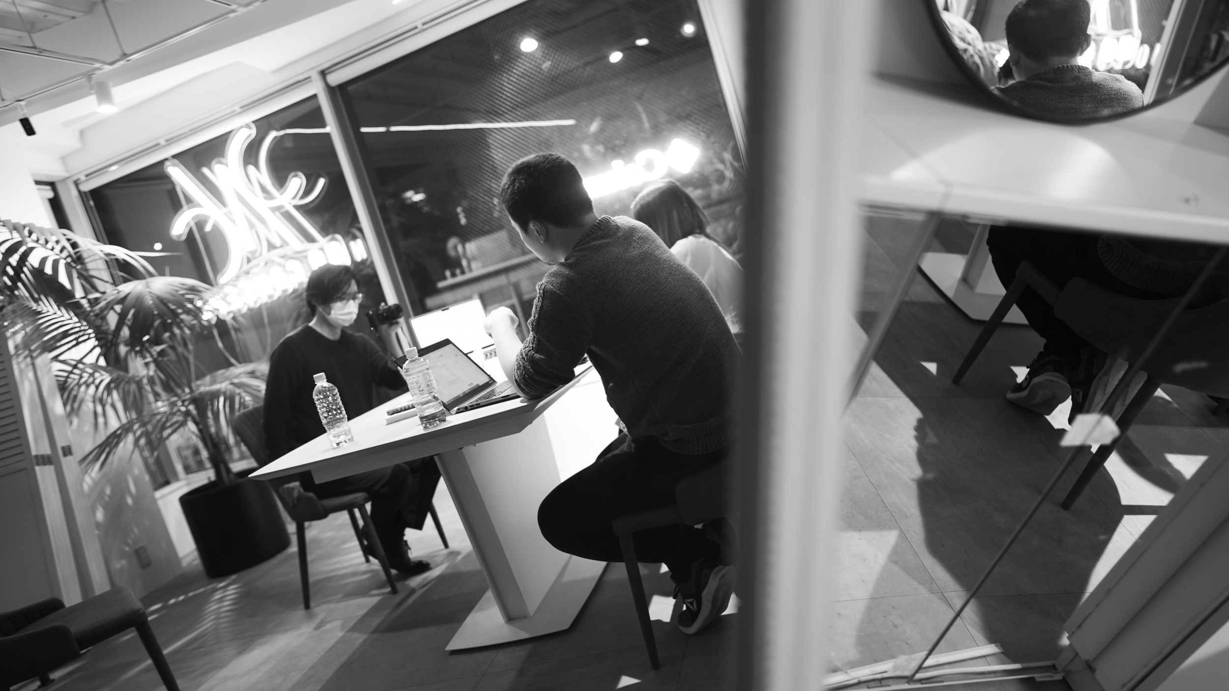 カフェの店内で人々が会話をしている様子。中央に男性が座ってパソコンを操作し、別の男性と話している。テーブルに水のボトルが置かれ、背景にネオンの看板が光っている。