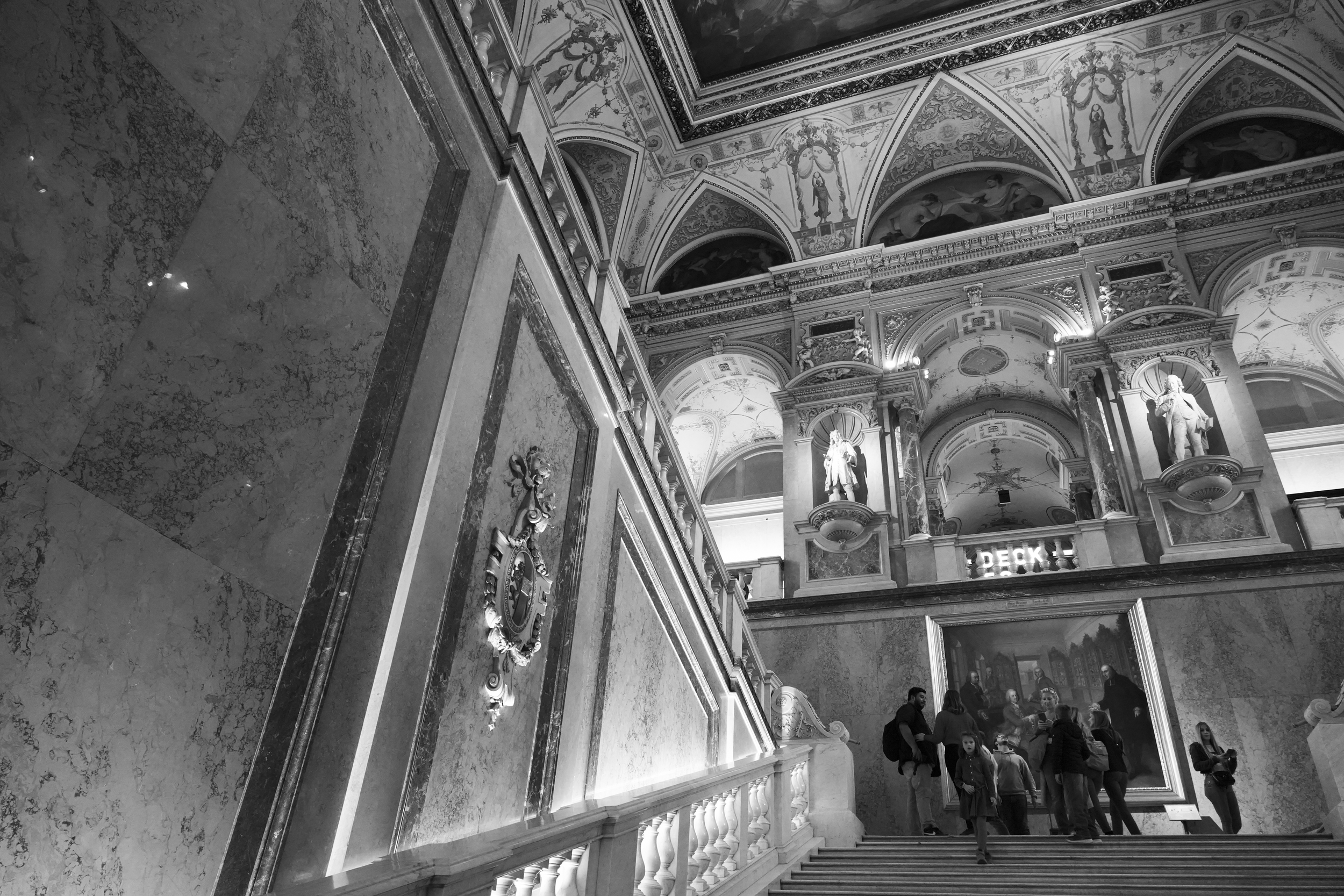 豪華な装飾が施された美術館の内装、階段の上に人々が集まり、絵画と彫刻が飾られている
