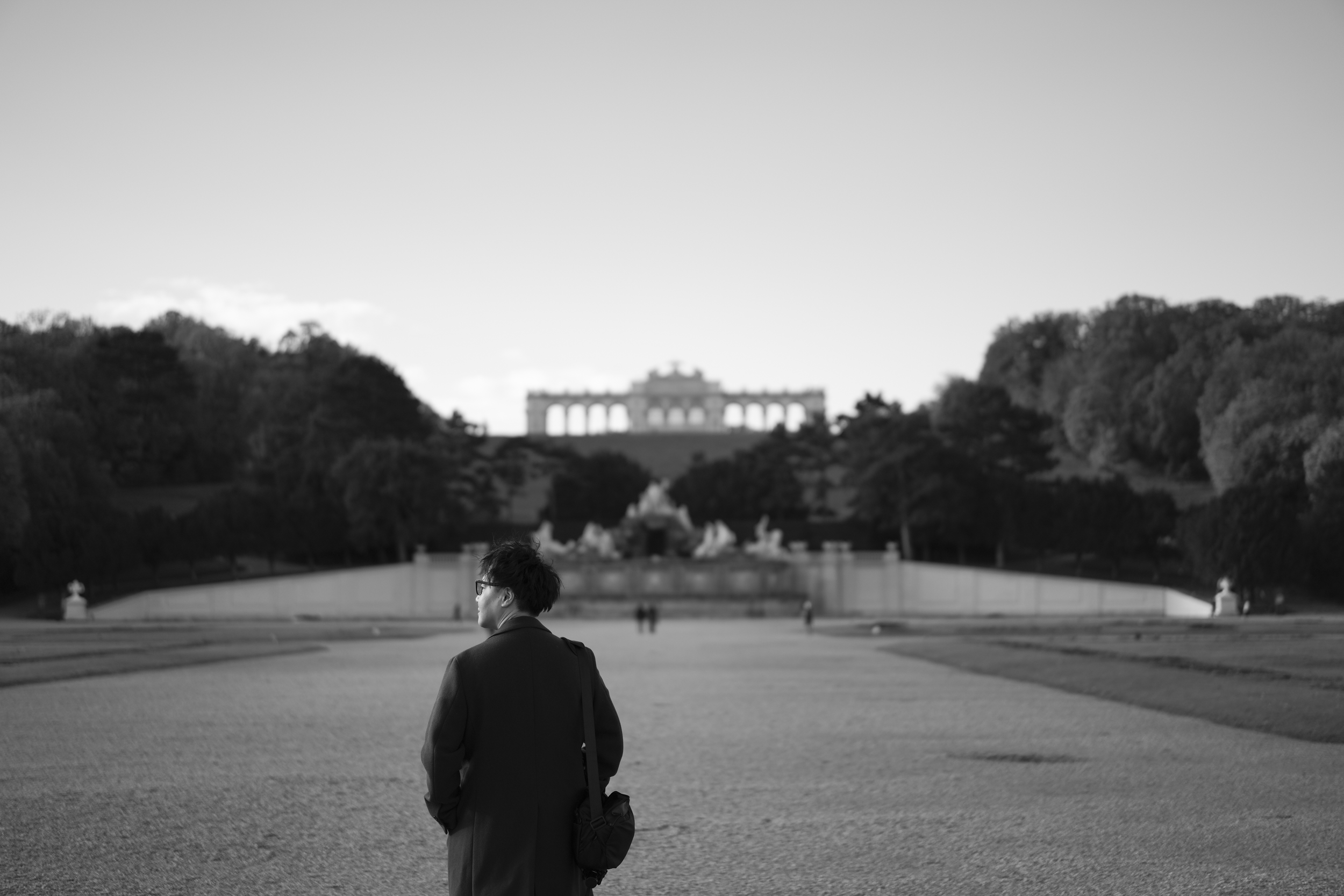 公園を歩く男性のシルエットと背景にアーチ型の建物と彫像が見える風景。