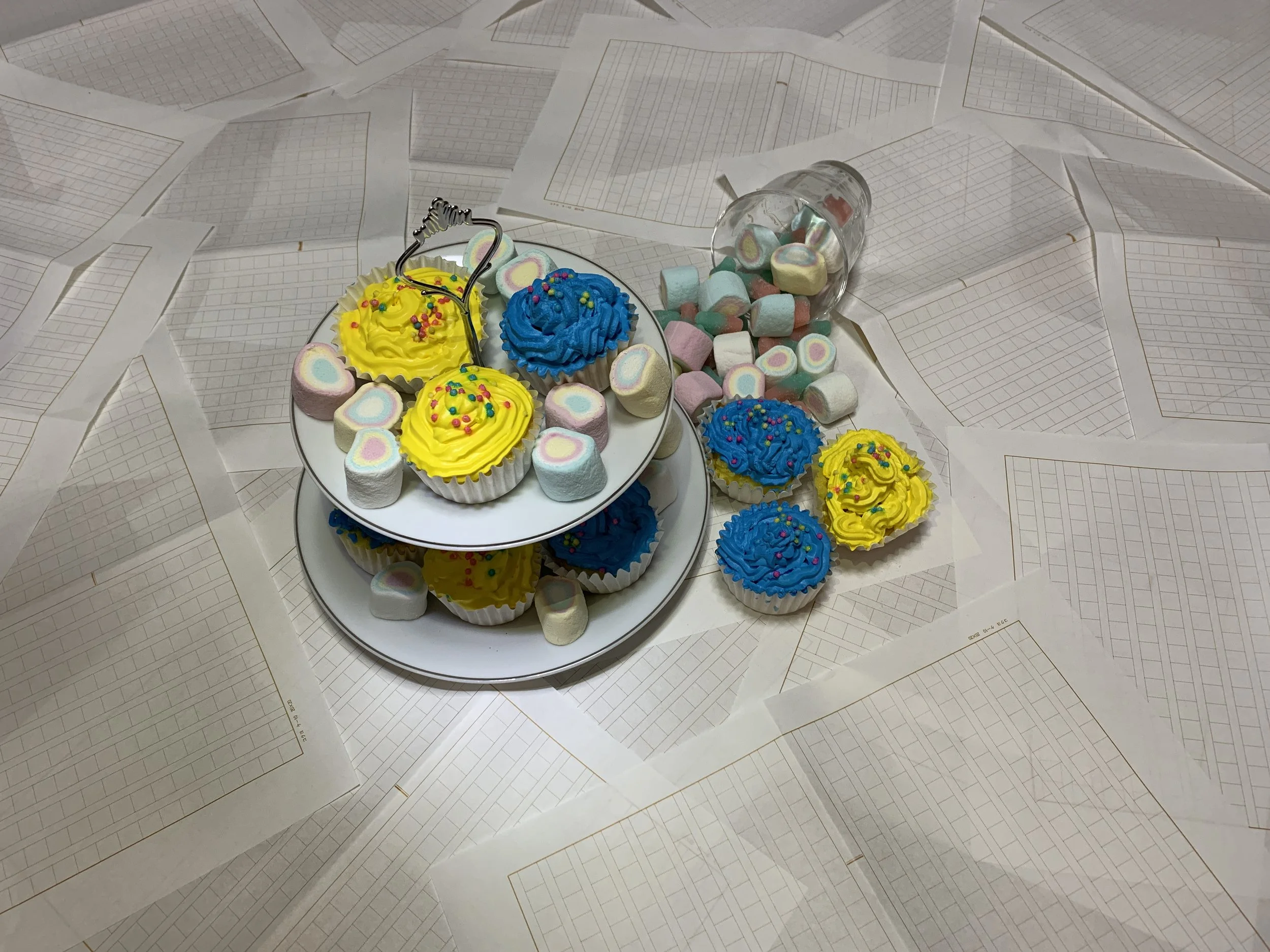 カラフルなマシュマロとカップケーキがプレートに乗っており、その周りに紙のノートや紙のシートが散らばっている