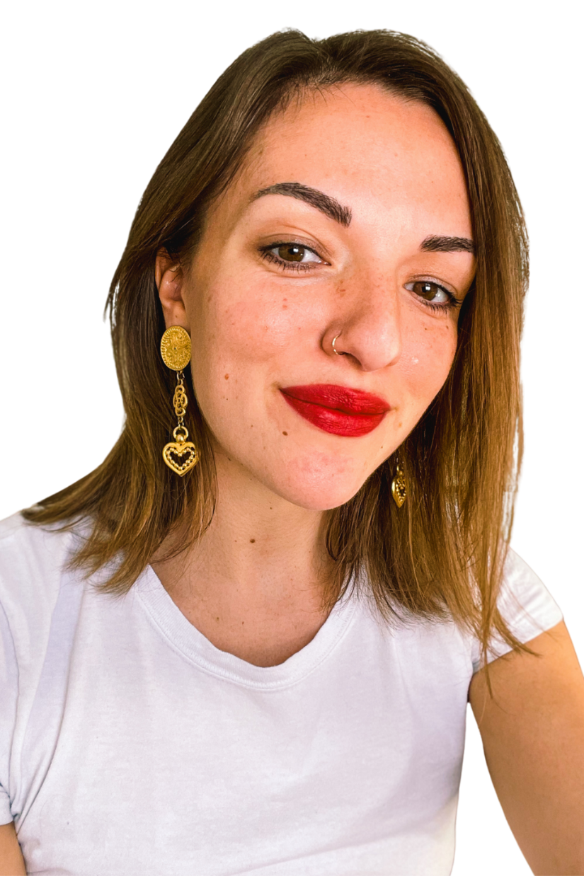 Close-up of a woman with shoulder-length brown hair, freckles, red lipstick, and gold earrings, wearing a white t-shirt, smiling at the camera.