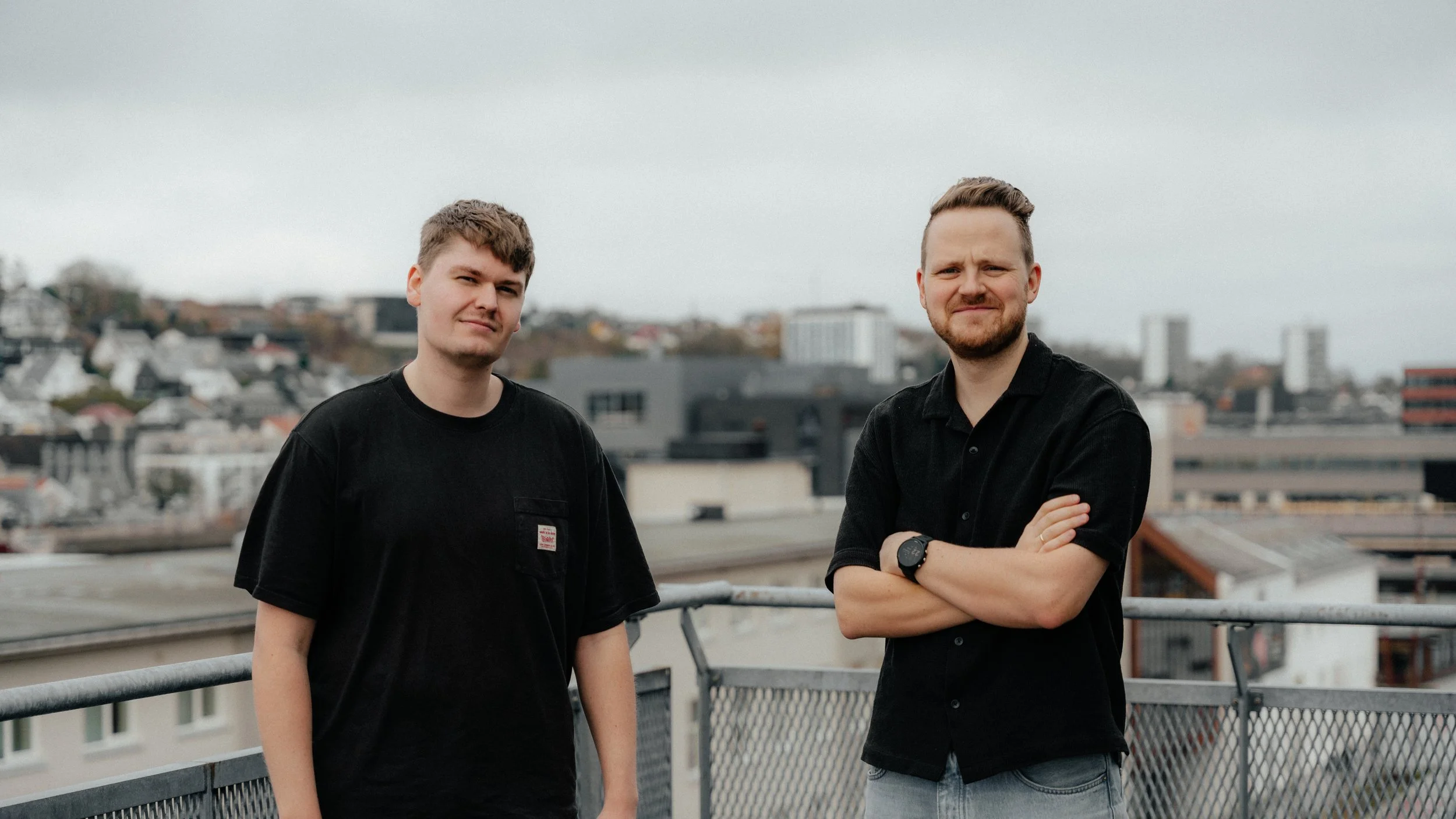 Two young men standing on a rooftop with a cityscape in the background, one with crossed arms and the other with a relaxed posture.