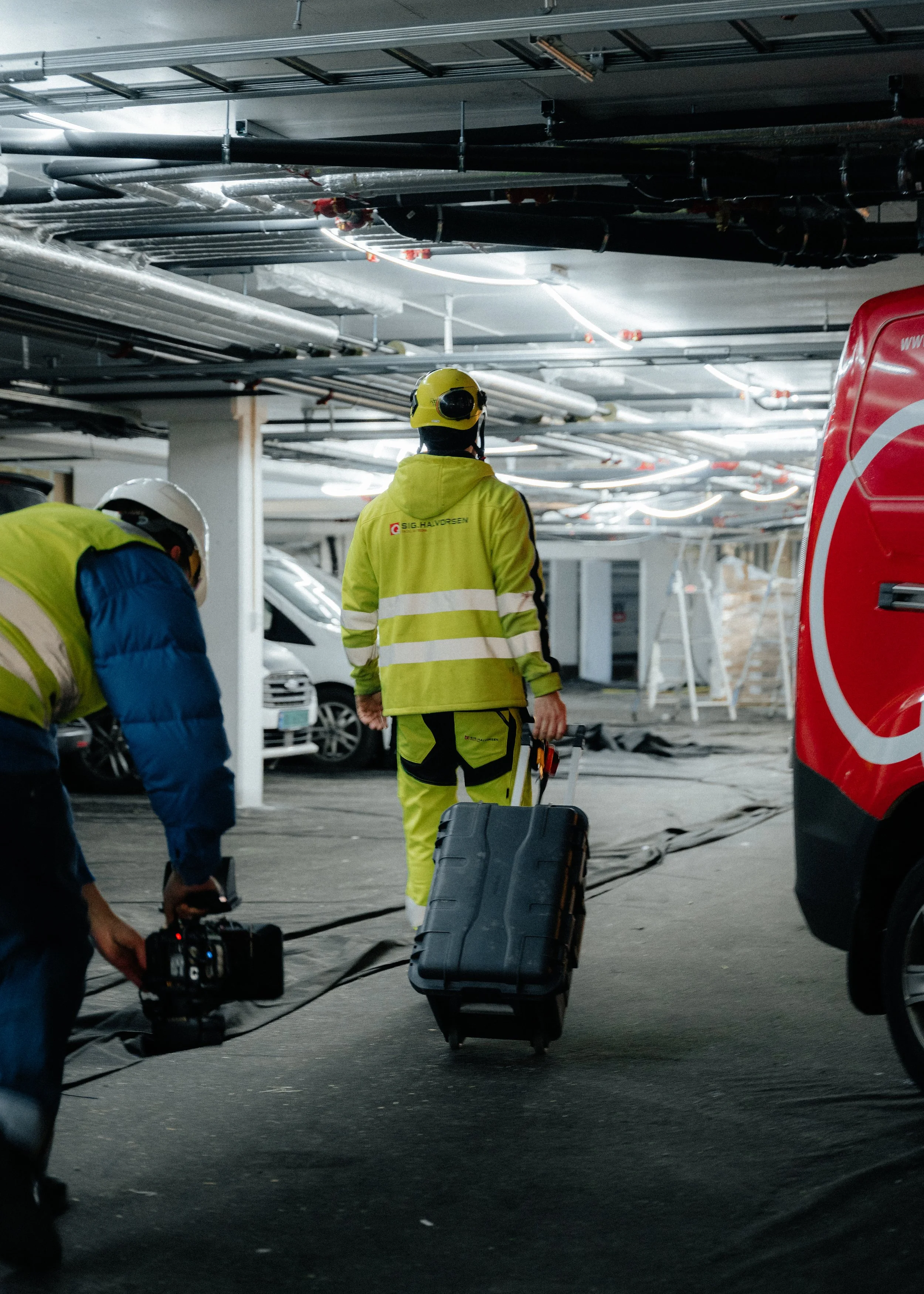 Workers in safety gear walking through an underground parking garage, one pulling a black toolbox on wheels, overhead pipes and vehicles in the background.