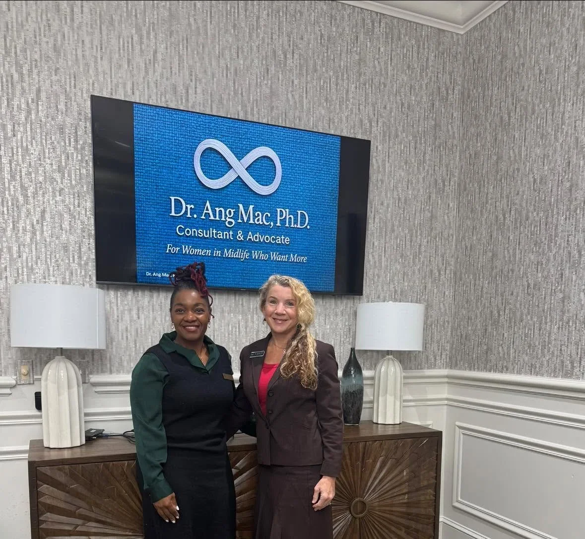 Two women pose together beneath a sign for Dr. Ang Mac, Ph.D., Consultant & Advocate for Women in Midlife Who Want More, highlighting a collaboration to empower women at Four Seasons in historic Virginia.