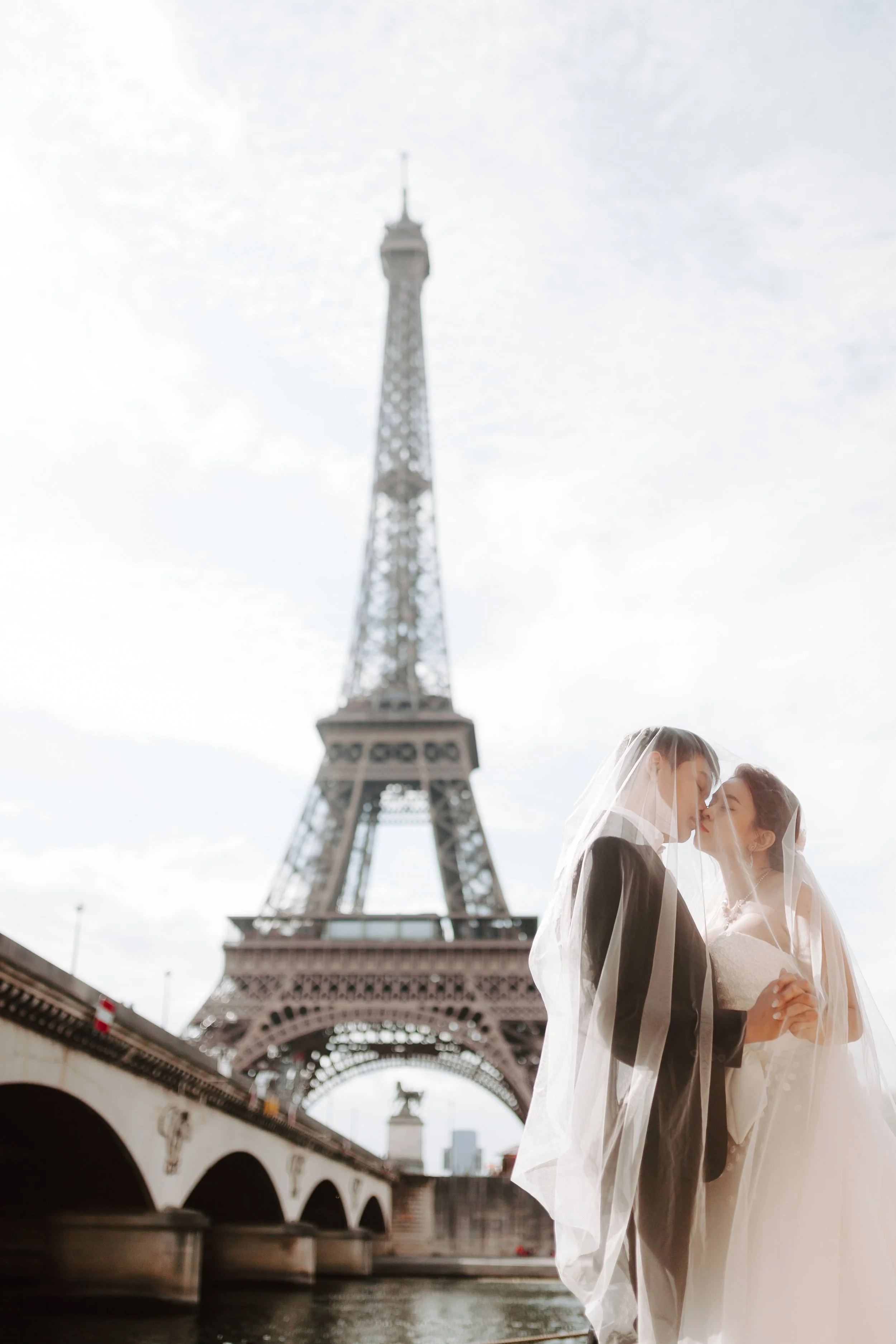 Two brides in wedding dresses and veils stand close to each other, touching noses, with the Eiffel Tower in Paris in the background.