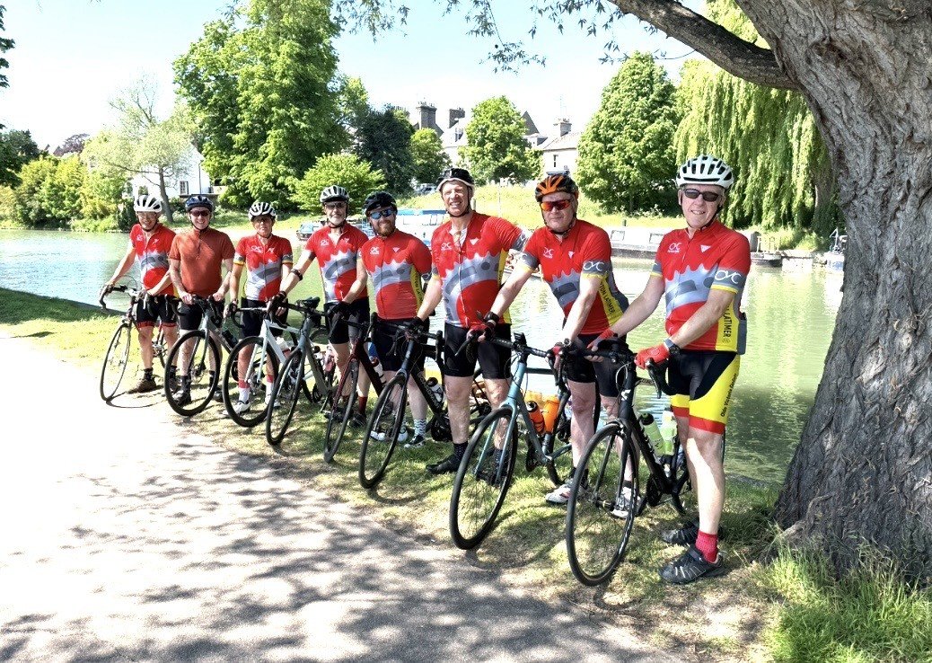 A group of seven cyclists in red cycling jerseys and helmets standing with bikes beside a lake, with green trees and a building in the background on a sunny day.