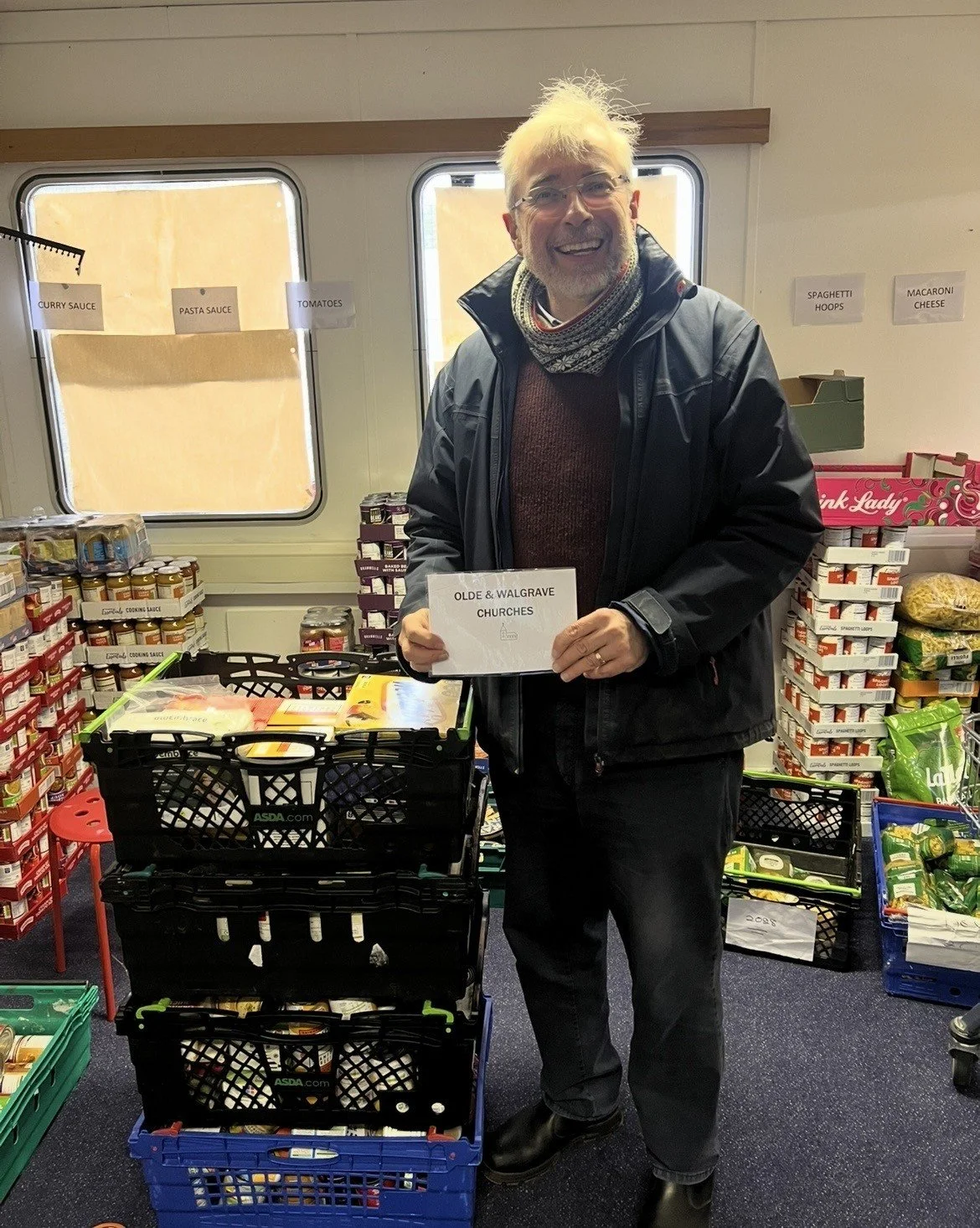 A man with white hair, glasses, and a beard smiling while holding a sign that reads 'OLDE & WALGRAVE CHURCHES' inside a store, with shelves stocked with canned goods and jars in the background.