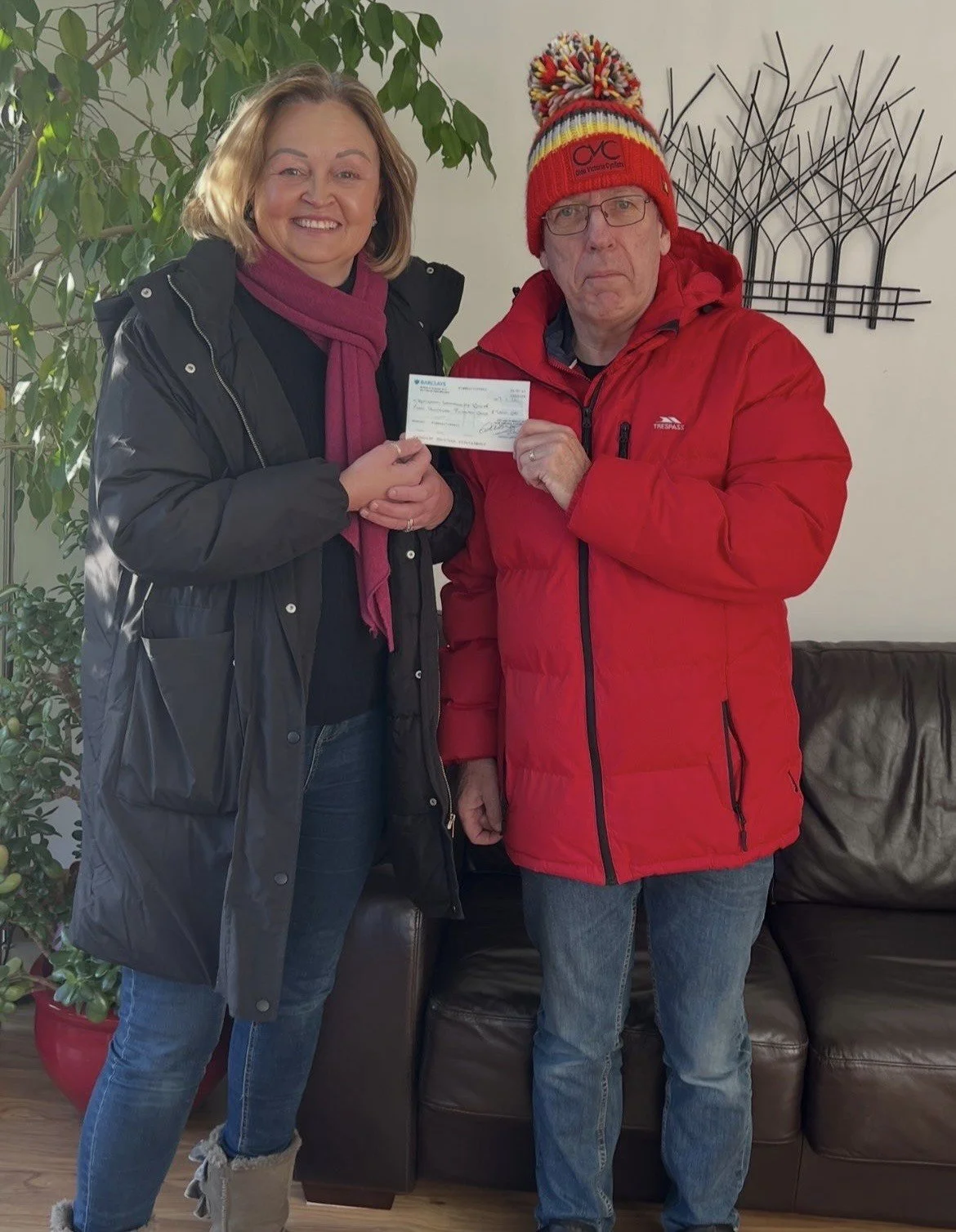 A woman and a man stand inside near a plant and a wall decoration. The woman is smiling and holding a check, while the man looks serious and is holding the check as well. Both are dressed warmly, with the man wearing a red jacket and a colorful knit hat.