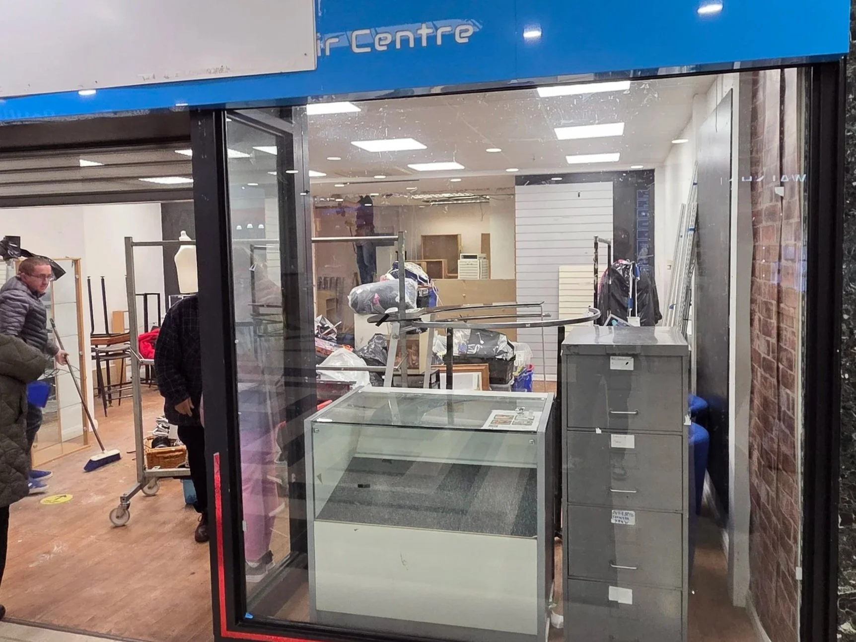 View inside a store or office through large glass window, showing cluttered space with desks, chairs, and people cleaning or organizing.