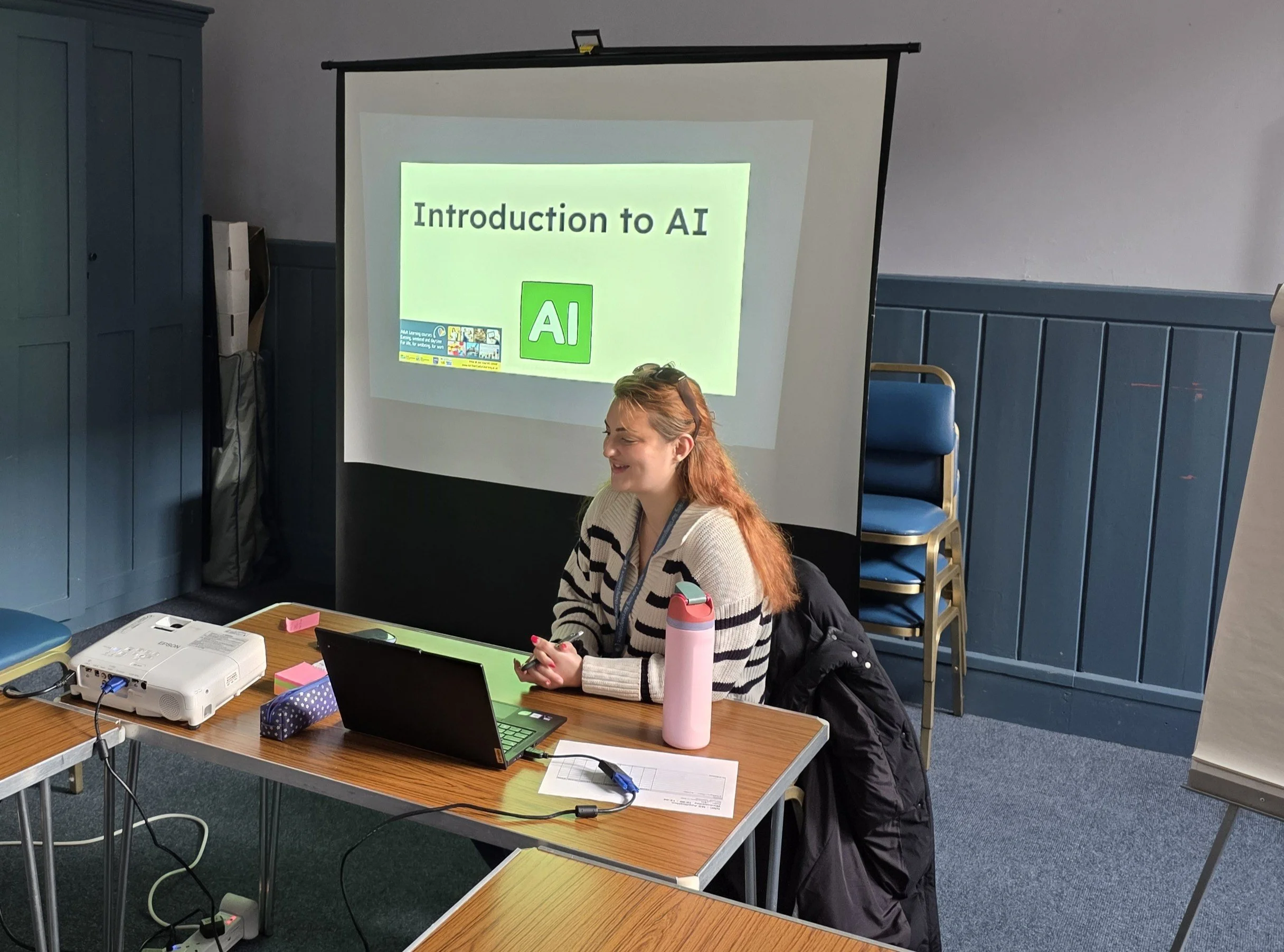 A woman sitting at a table looks to her side, smiling, with a presentation slide in the background labeled 'Introduction to AI'. The table has a laptop, pink water bottle, some sticky notes, and papers. A projector is connected to the laptop.