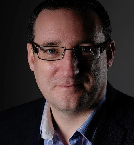 Close-up portrait of a man wearing glasses, dark suit, and light blue shirt, with a serious expression on a dark background.