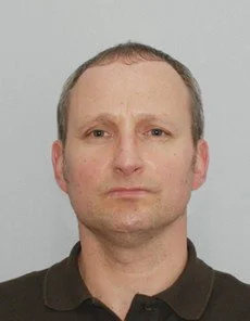 Headshot of a man with short hair and a serious expression wearing a dark collared shirt against a light background.