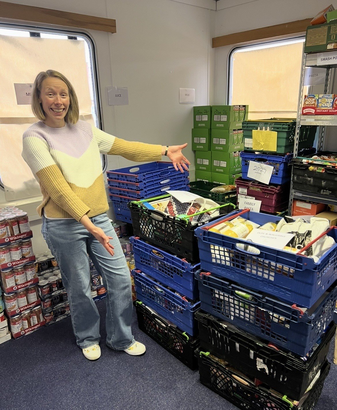 A woman is standing in a room, smiling, and pointing towards stacked plastic crates filled with various food items. The crates are on the floor, and there are shelves and windows behind her, with signs and labels indicating different food categories.