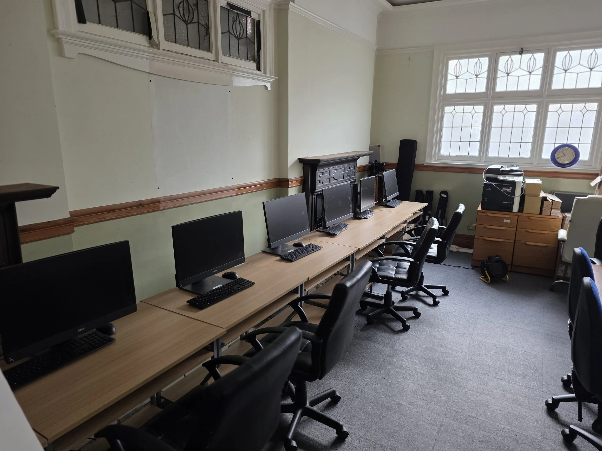 A room with a row of computers on desks along the wall, black office chairs, a large window letting in natural light, and a printer on a wooden cabinet.