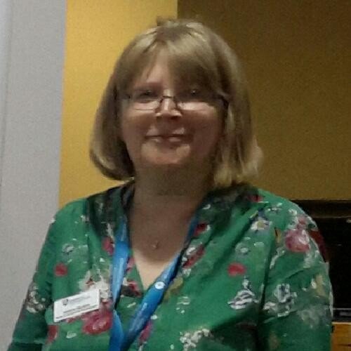 A woman smiling with glasses, wearing a green floral shirt and a blue lanyard, standing in front of a yellow and gray wall.