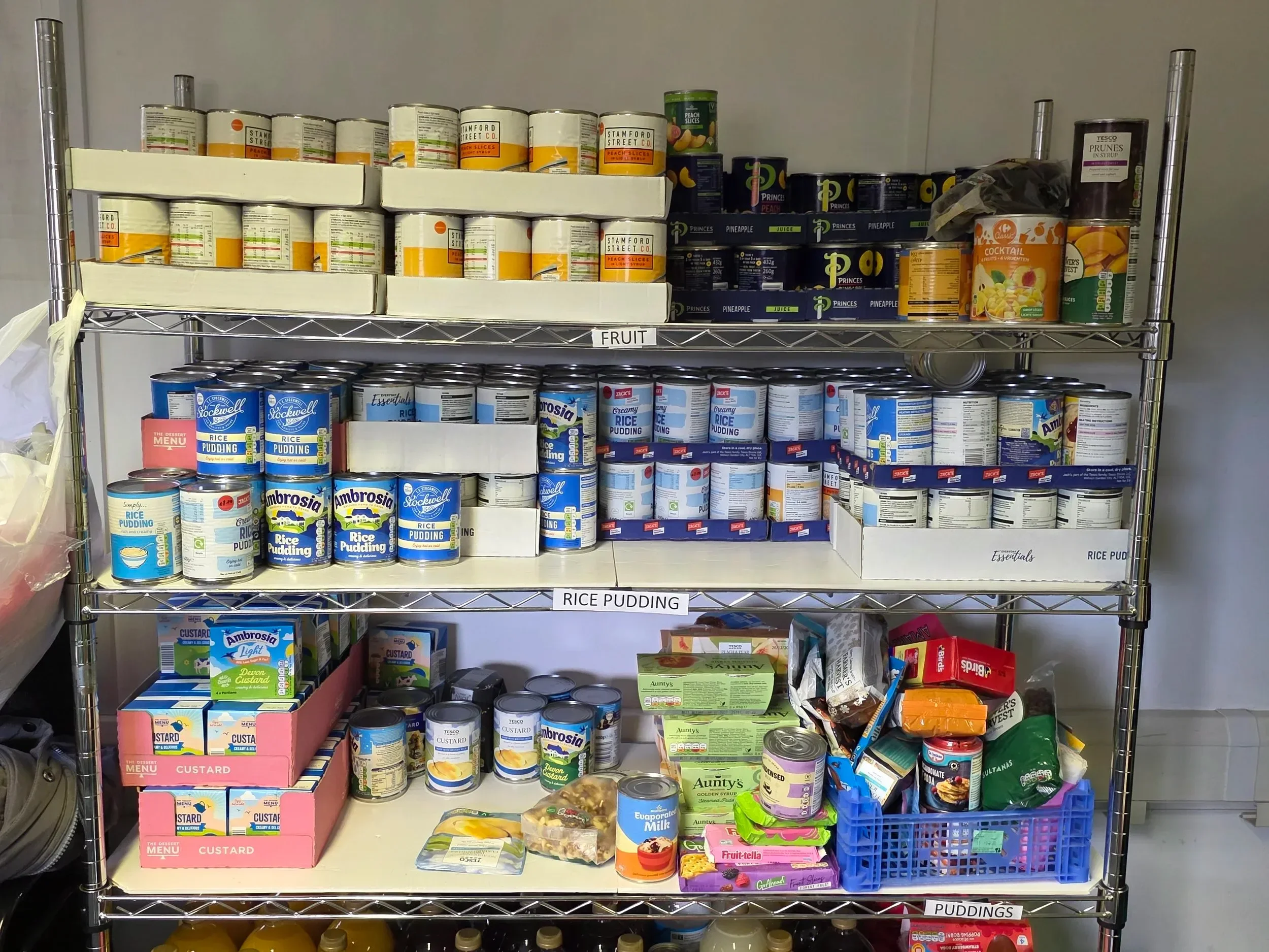 Shelves stocked with canned goods, including rice pudding, custard, canned fruit, and other pantry items, arranged in sections labeled "FRUIT," "RICE PUDDING," and "PUDDINGS."