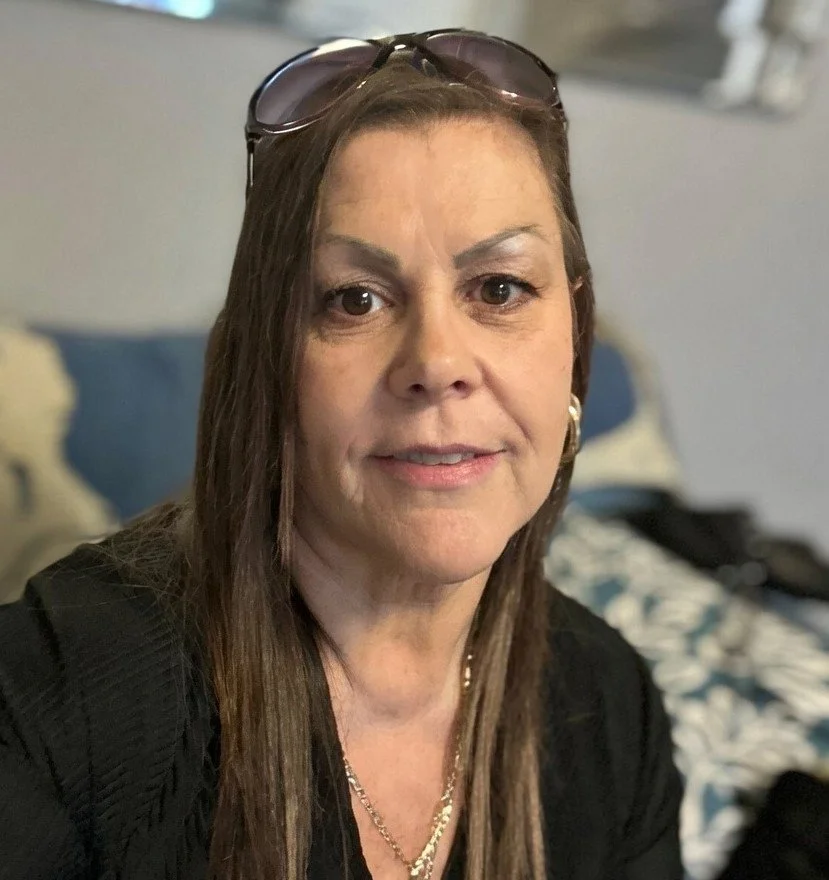A woman with long brown hair wearing sunglasses on her head, a black top, and jewelry, sitting on a bed with a blue headboard and patterned bedding in the background.