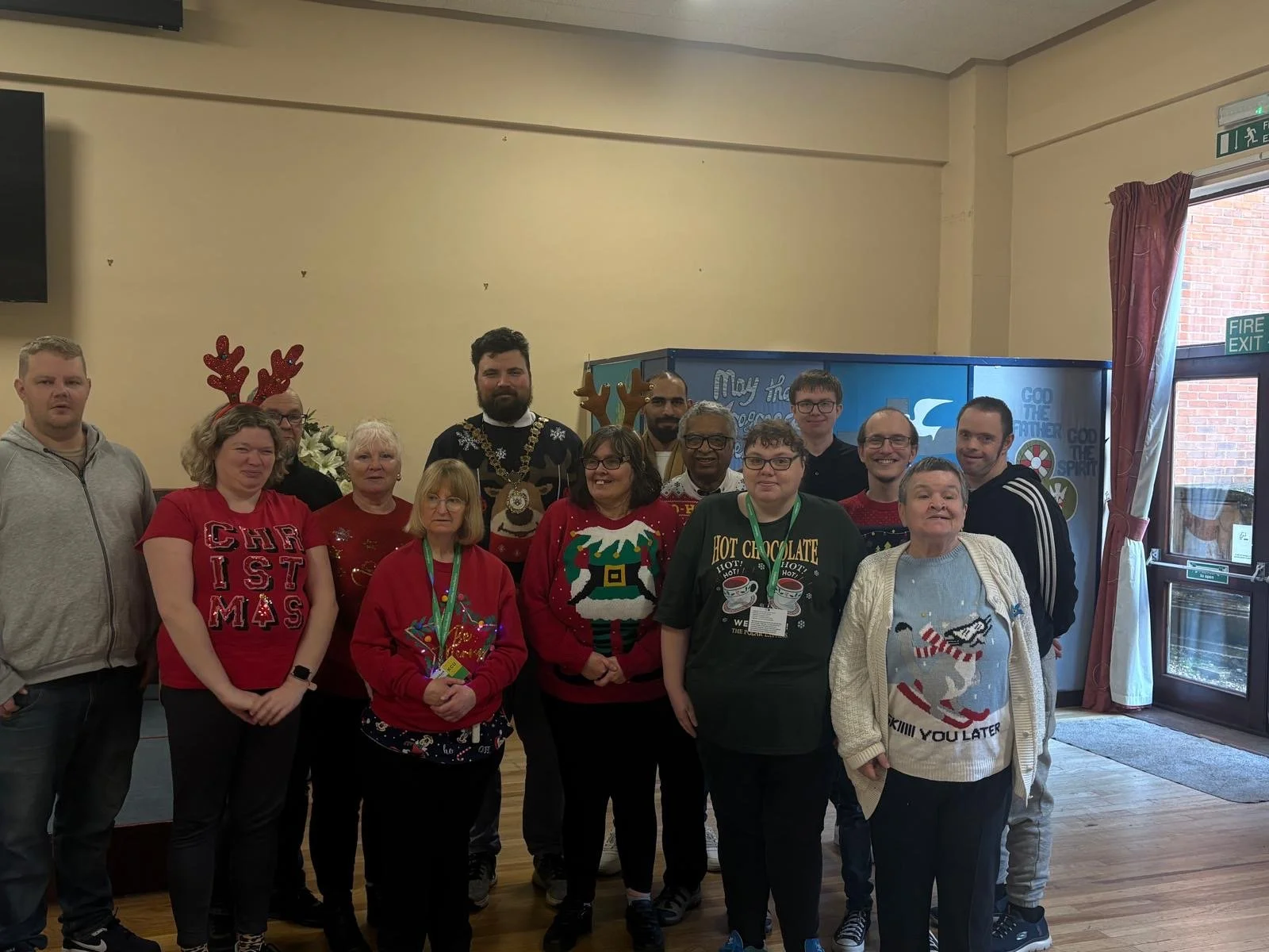 Group of people celebrating Christmas indoors, some wearing festive sweaters and reindeer antler headbands, in a decorated room with a blue game machine in the background and a window with curtains on the right.