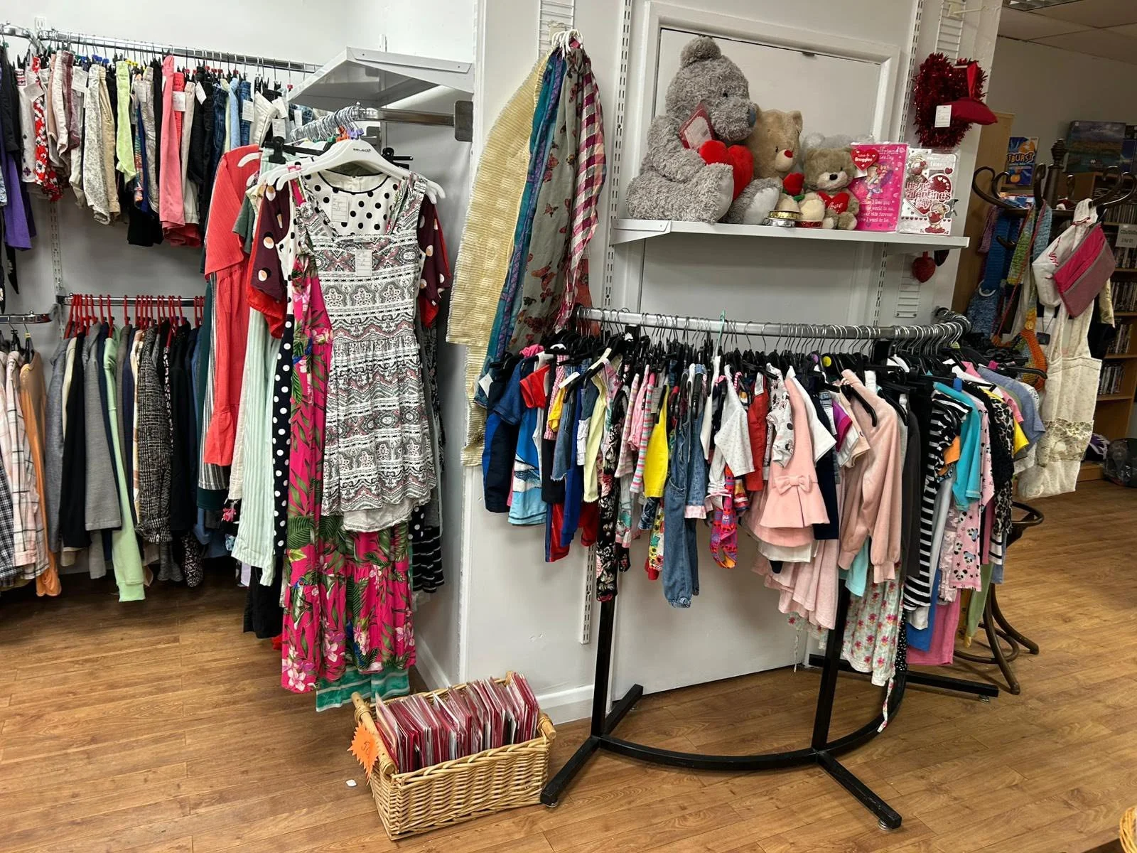 Clothing display in thrift store with a rack of children's clothes, a basket of magazines or books, and shelves with stuffed animals, toys, and holiday decorations.