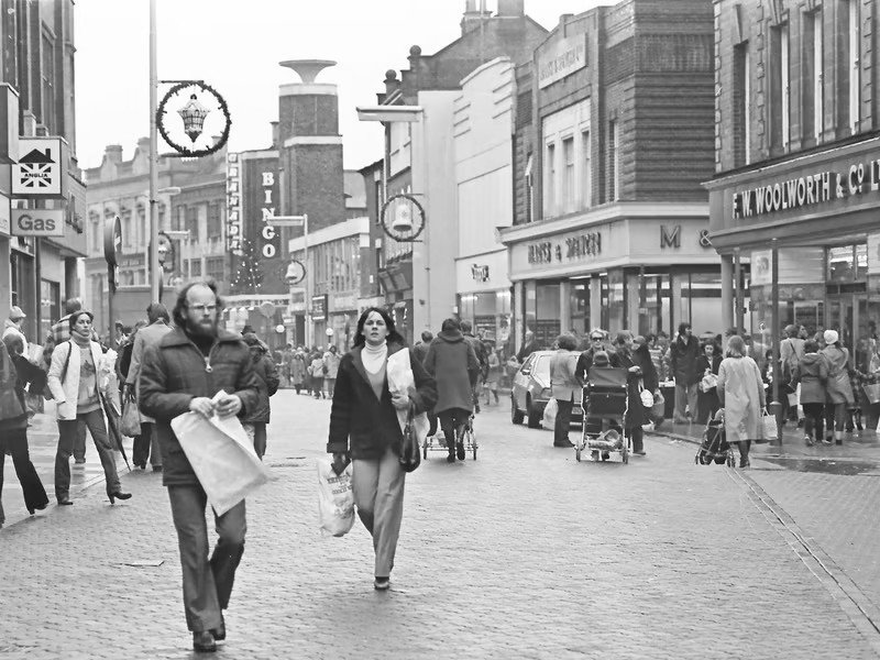 A busy city street with people walking, shopping, and pushing strollers in black and white.