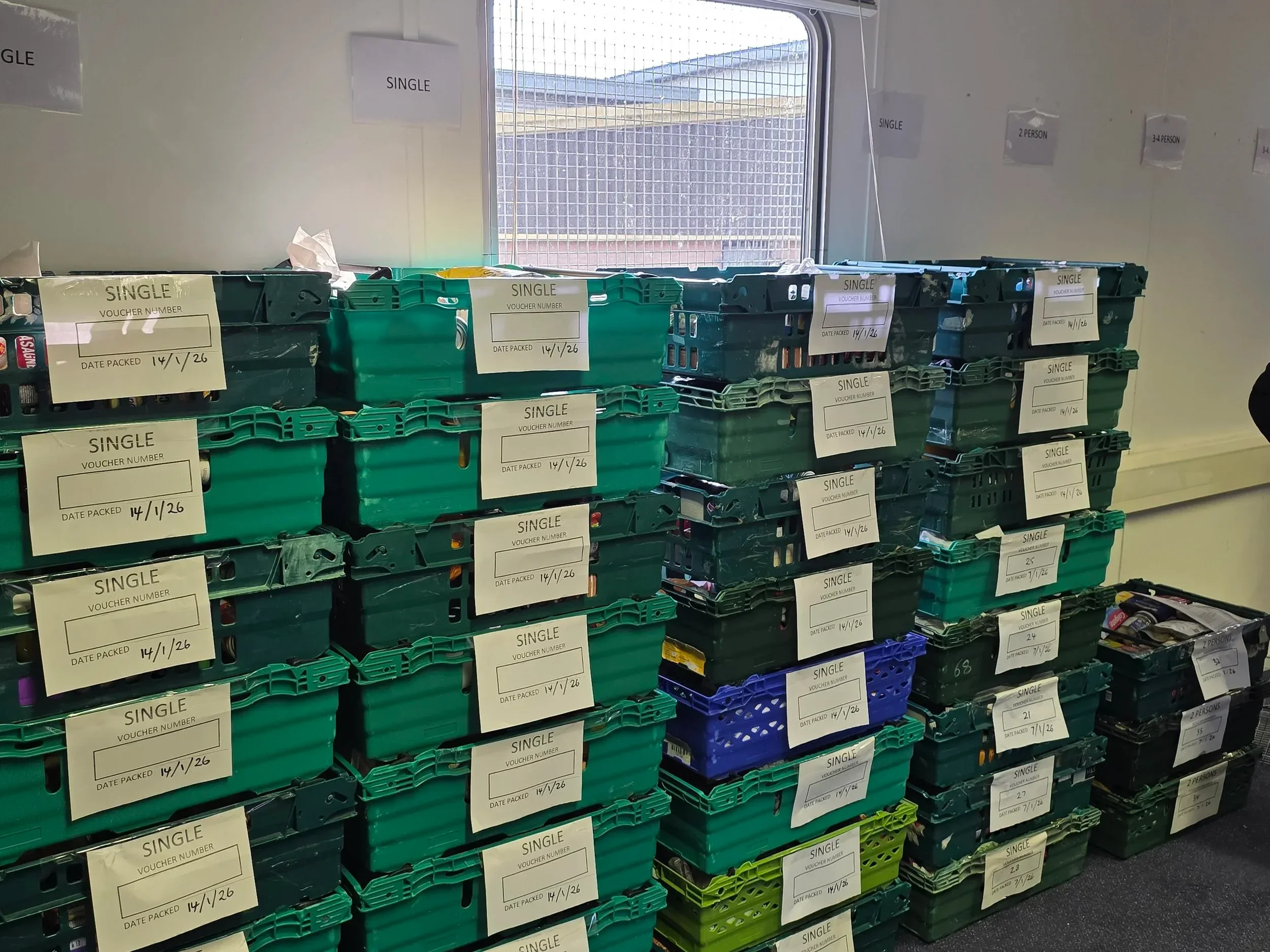 Stacked green and black plastic crates with white labels indicating 'SINGLE' and the date '14/1/26', placed in front of a window in a room that appears to be a storage or distribution area.