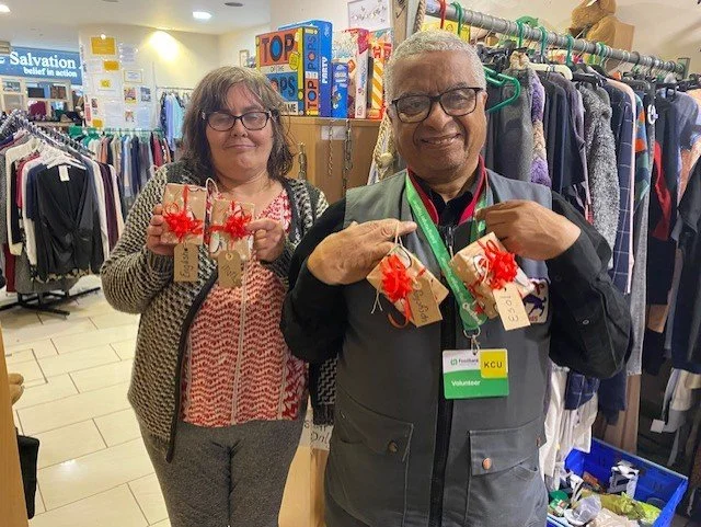 Two volunteers holding wrapped gifts in a thrift store with clothing racks in background.